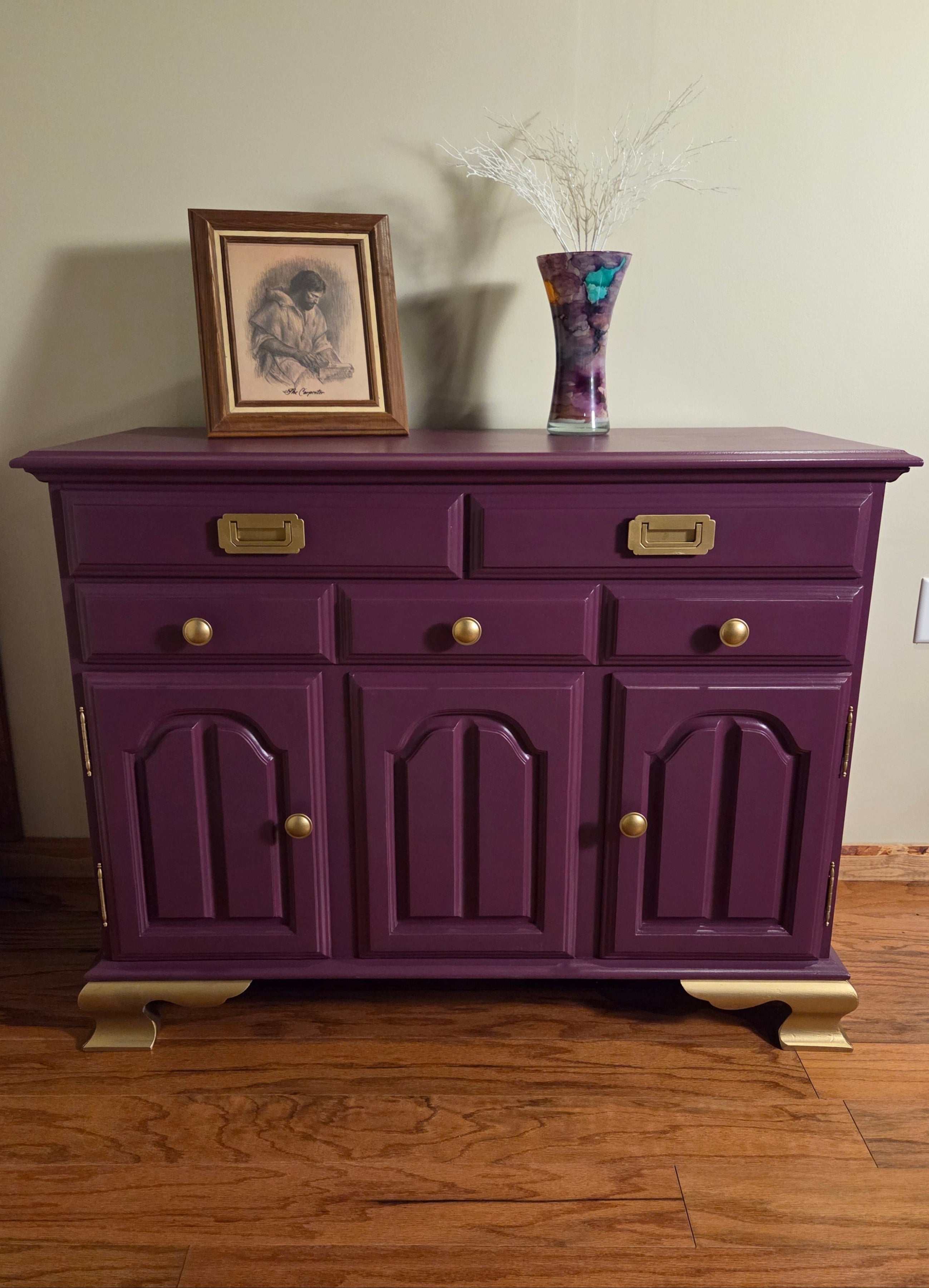Purple dresser with gold knobs on a wooden floor against a beige wall.