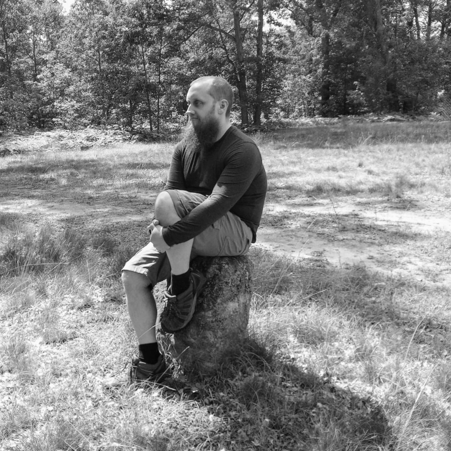 A man with a beard sits on a bolder in a grassy field with trees in the background, shown in black and white.